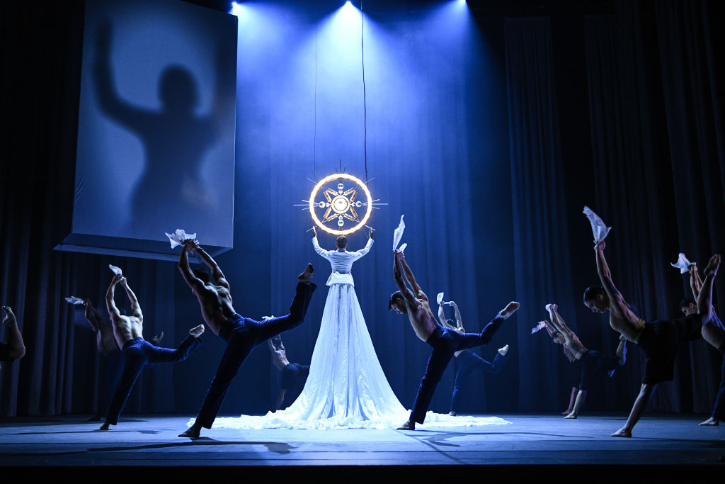 A person in white raises her arms to a glowing circle while dancers move around her.