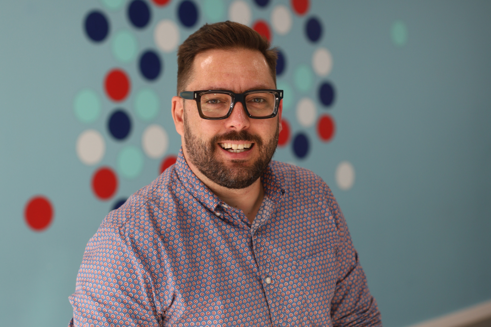 Man in a button-down smiles at camera in front of a blue wall with blue, white and red spots.