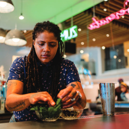 A person puts herb leaves in a bowl, with a colorful restaurant in the background.
