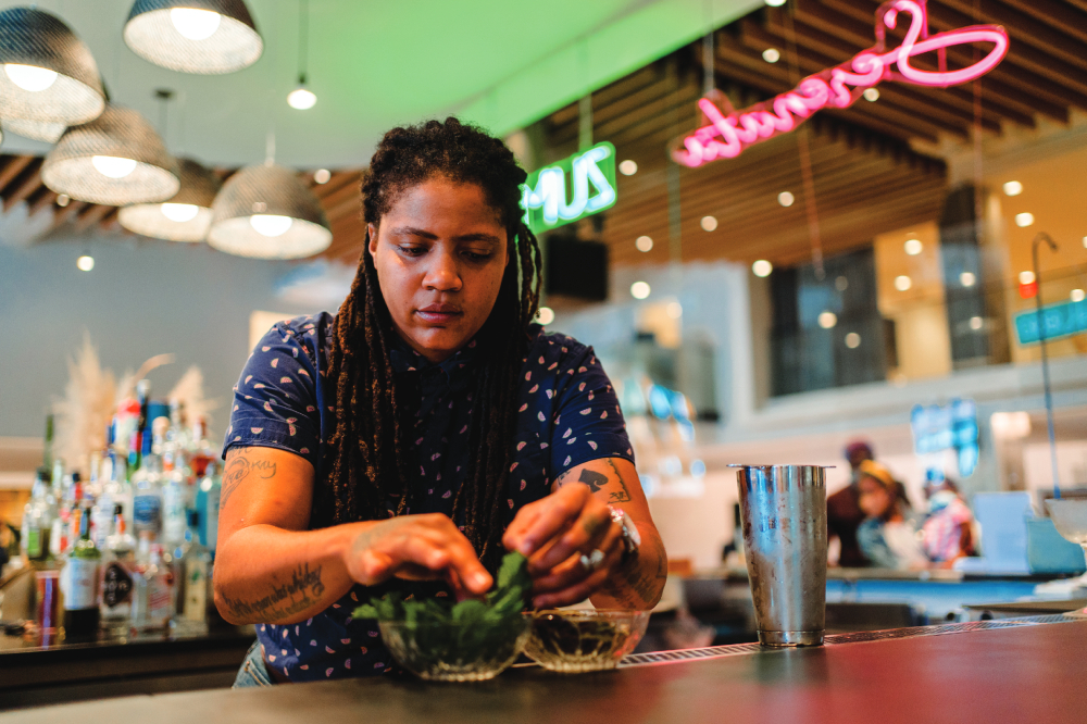 A person puts herb leaves in a bowl, with a colorful restaurant in the background.