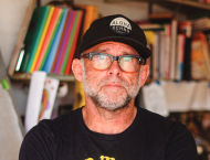 A man in a black t-shirt and baseball cap stands in front of a bookshelf.