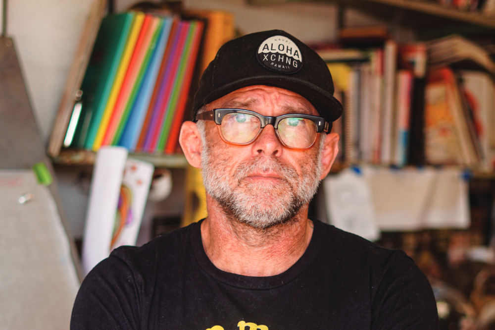 A man in a black t-shirt and baseball cap stands in front of a bookshelf.