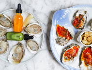 Oysters on the half shell sit in a circular pattern on a plate. Another plate serves oysters or another mollusk with red sauce.