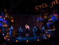A person raises her hand on stage, with others dancing around her.