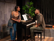 One person in jeans and a shawl leans on a piano, looking at the person sitting on the piano bench.