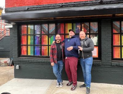 Three men stand in front of a building with pride flags in every window.