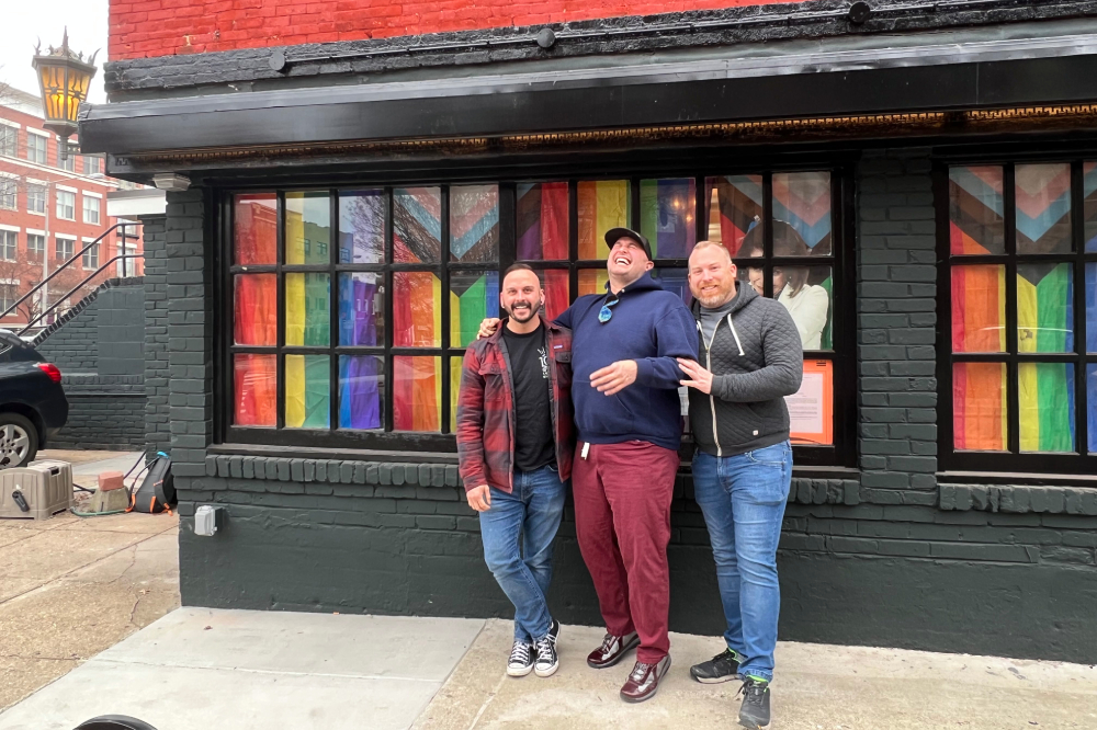 Three men stand in front of a building with pride flags in every window.