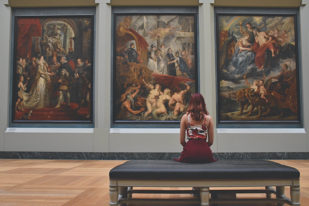 A person sits on a bench, looking at paintings in a museum.