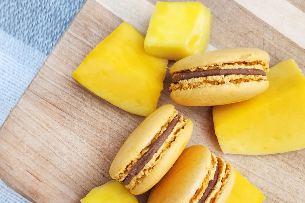 Three yellow macarons sit on a cutting board next to pieces of mango.