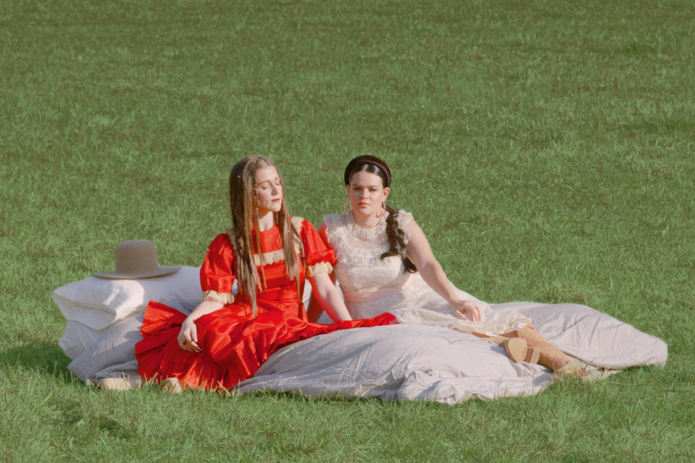 One person in a red dress and one in a light pink dress sit next to each other in the grass.