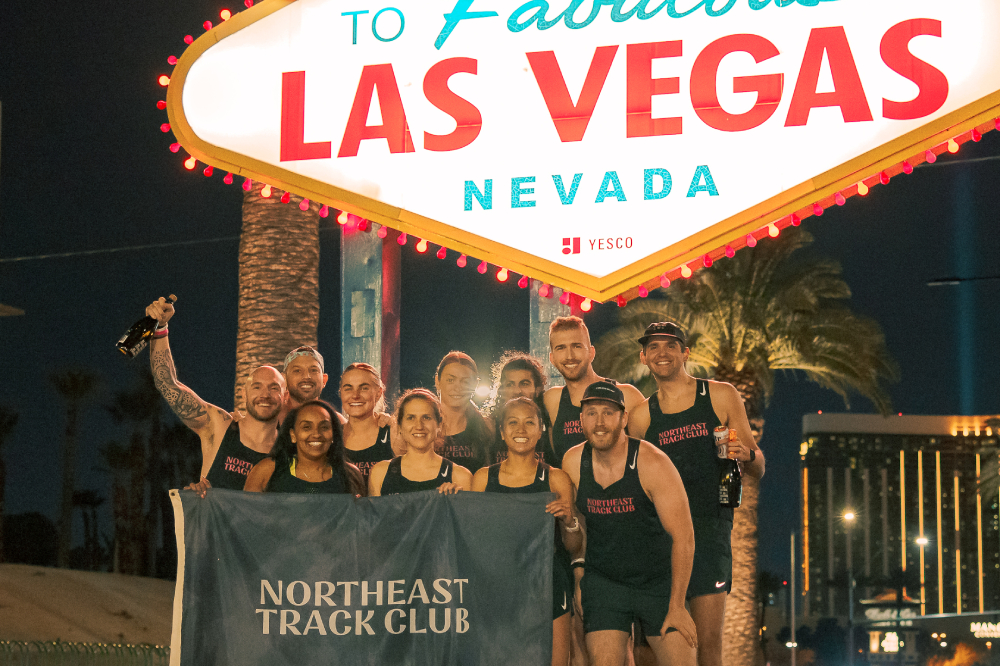 A group of people hold a Northeast Track Club flag in front of a Las Vegas sign.