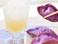 Next to a glass with a pale orange liquid, two purple dumplings sit on a plate and a pair of chopsticks pick up a third.