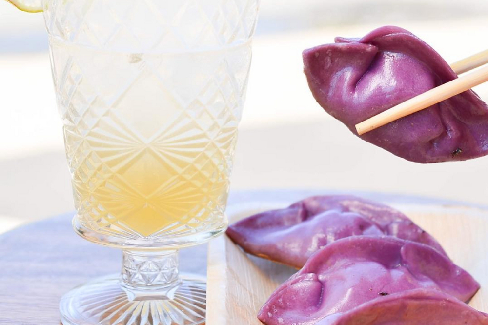 Next to a glass with a pale orange liquid, two purple dumplings sit on a plate and a pair of chopsticks pick up a third.