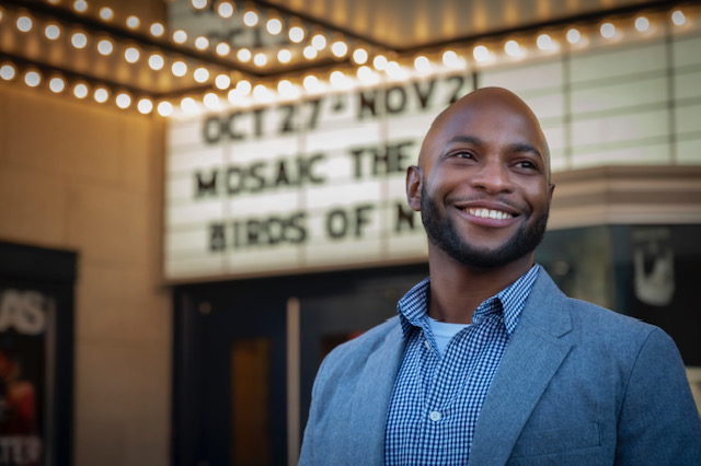 A man in a blue shirt looks to the right in front of Mosaic Theater.
