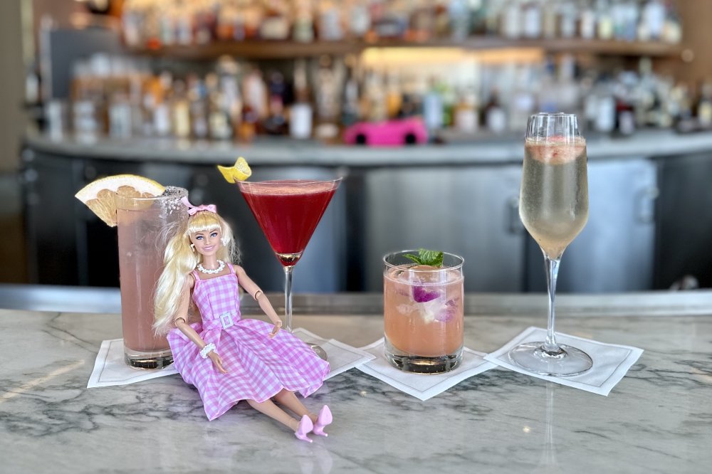A Barbie doll sits among pink drinks on a bar counter.